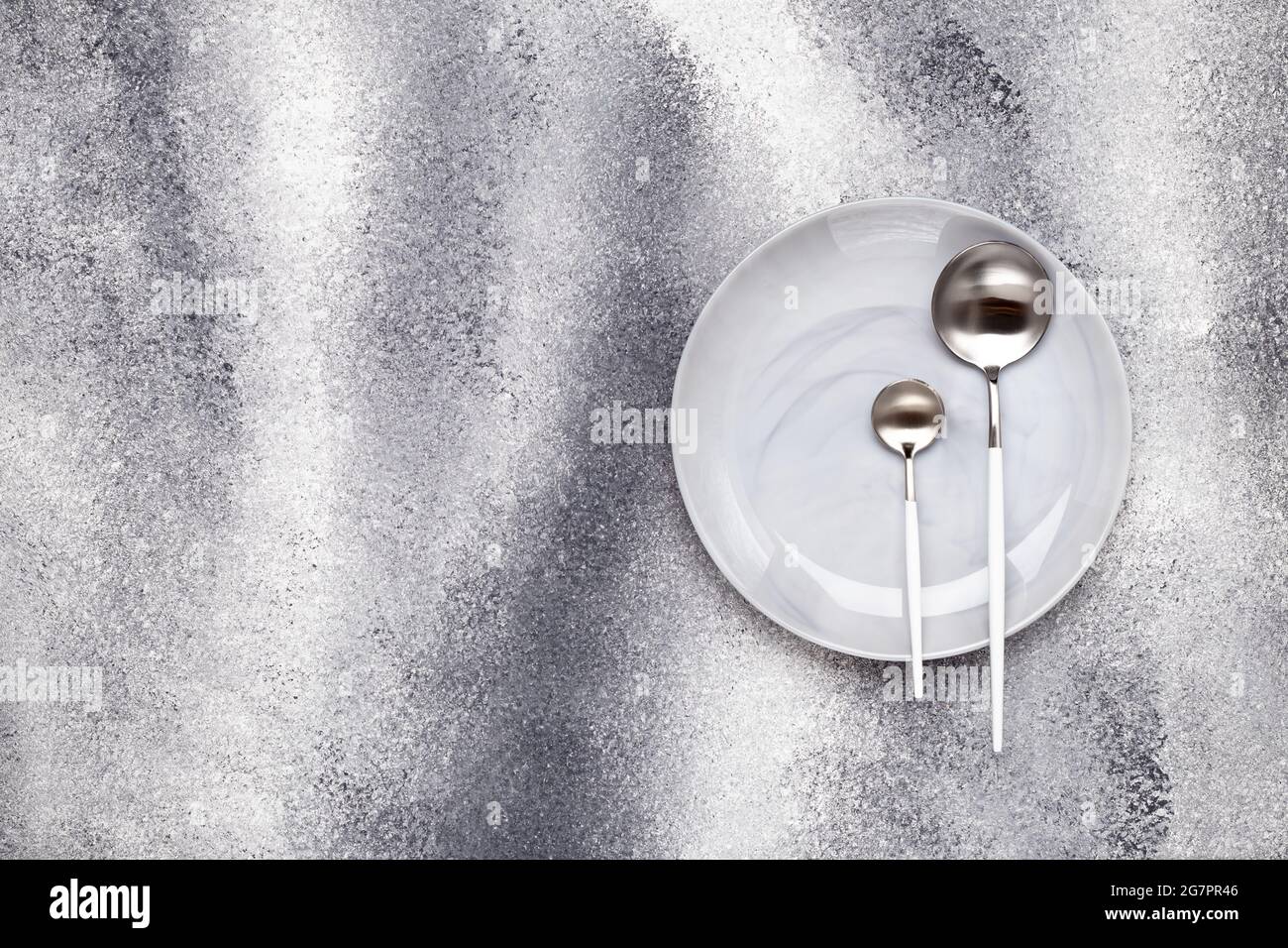 Svuotare la piastra in ceramica pulita e il cucchiaino da tè non utilizzato con il cucchiaio da tavola. Stoviglie e posate su un tavolo grigio a grunge, che serve concetto. Menu di progettazione monocromatica. Foto Stock