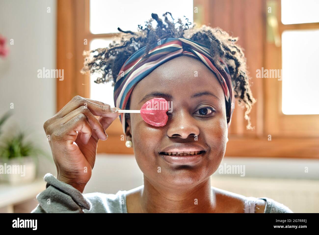 Closeup divertente ritratto di una giovane donna nera con acconciatura afro scherzare con un lollipop indoor a casa, ridendo. Concetto di stile di vita. Foto Stock