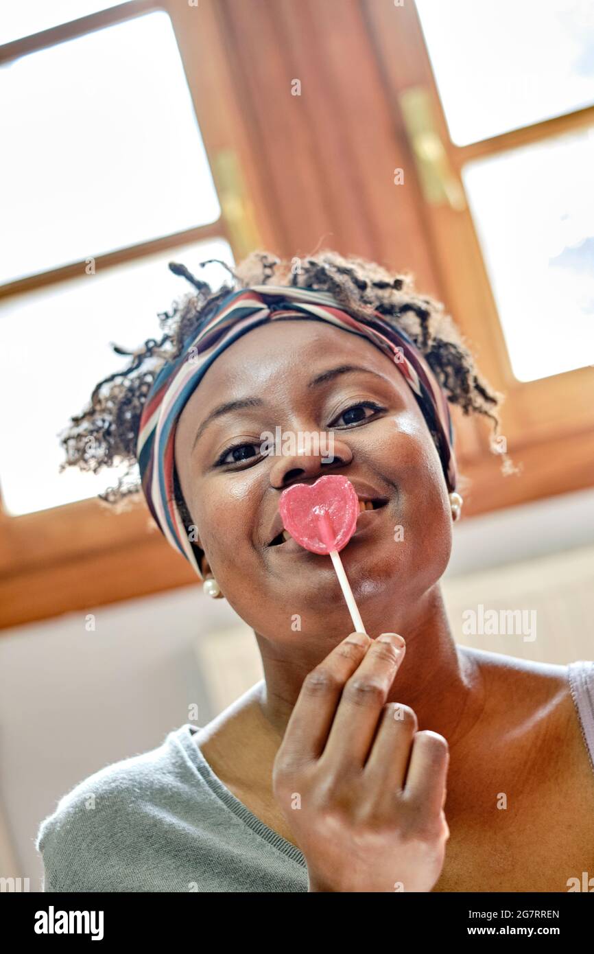 Closeup divertente ritratto di una giovane donna nera con acconciatura afro scherzare con un lollipop indoor a casa, ridendo. Concetto di stile di vita. Foto Stock