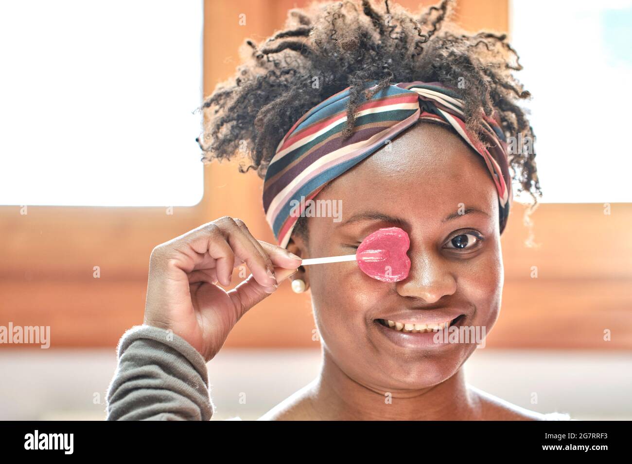 Closeup divertente ritratto di una giovane donna nera con acconciatura afro scherzare con un lollipop indoor a casa, ridendo. Concetto di stile di vita. Foto Stock