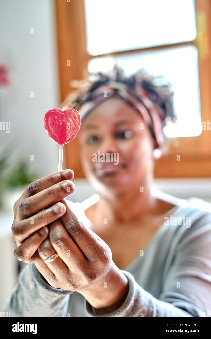 Ritratto closeup di una giovane donna nera con acconciatura afro che scherza con un lecca in casa. Concetto di stile di vita. Foto Stock