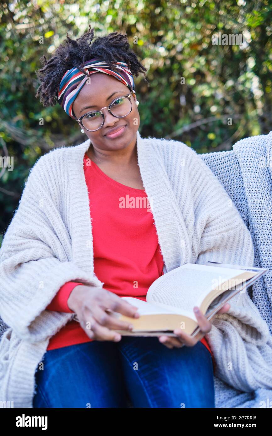 Ritratto di una giovane donna nera con acconciatura e occhiali afro che legge un libro seduto su una vecchia panchina giardino. Concetto di stile di vita. Foto Stock