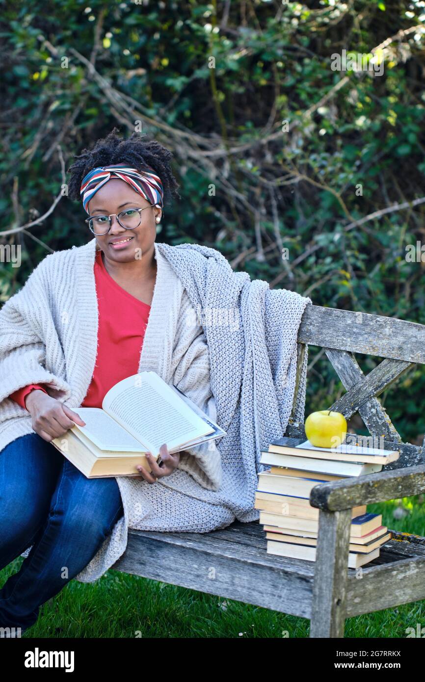 Ritratto di una giovane donna nera con acconciatura e occhiali afro che legge un libro seduto su una vecchia panchina giardino con un mucchio di libri. Concetto di stile di vita. Foto Stock