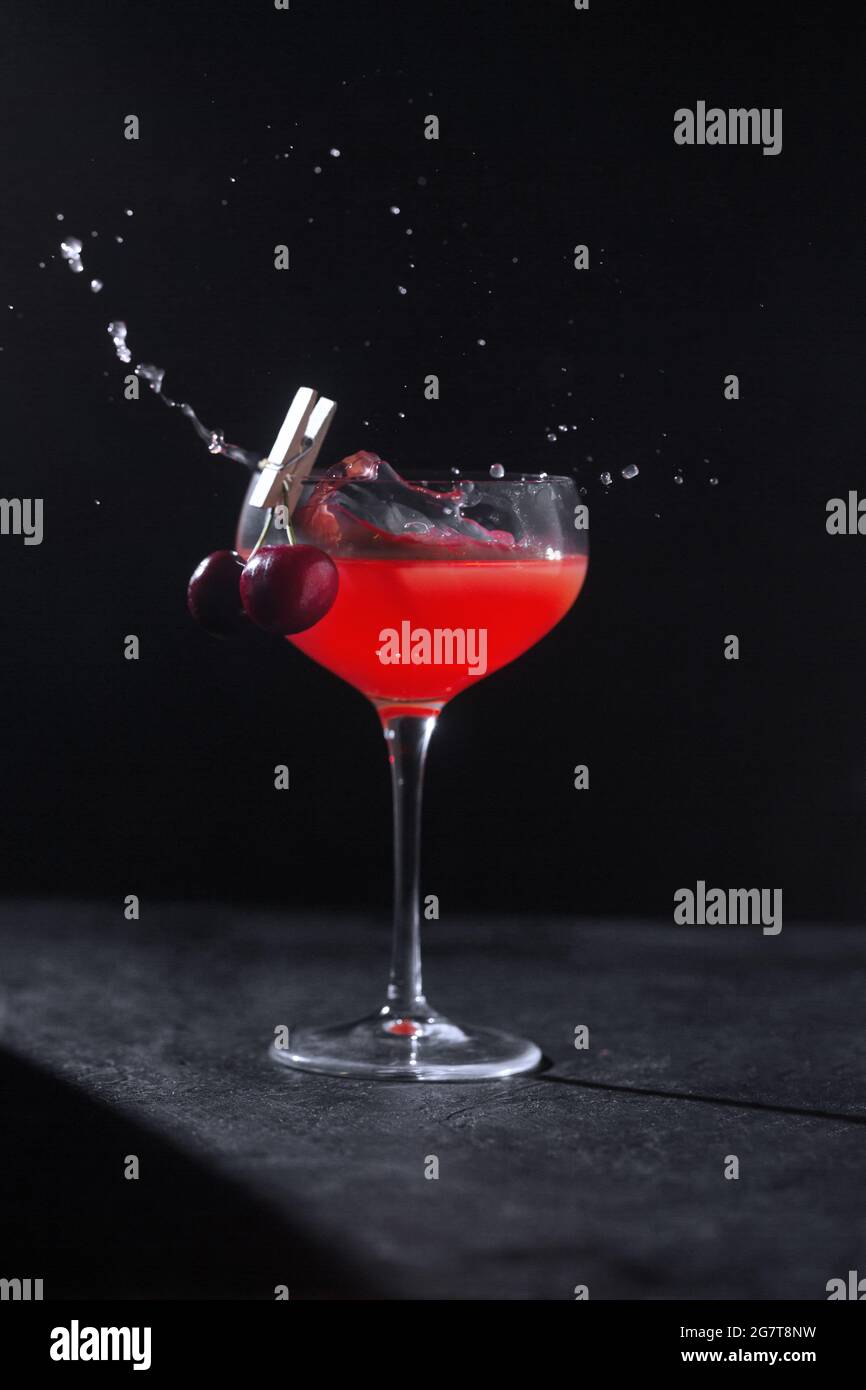 Un bicchiere con un cocktail alcolico fresco realizzato da un barman decorato con ciliegie su uno stendibiancheria e spruzzi. Tasto basso Foto Stock