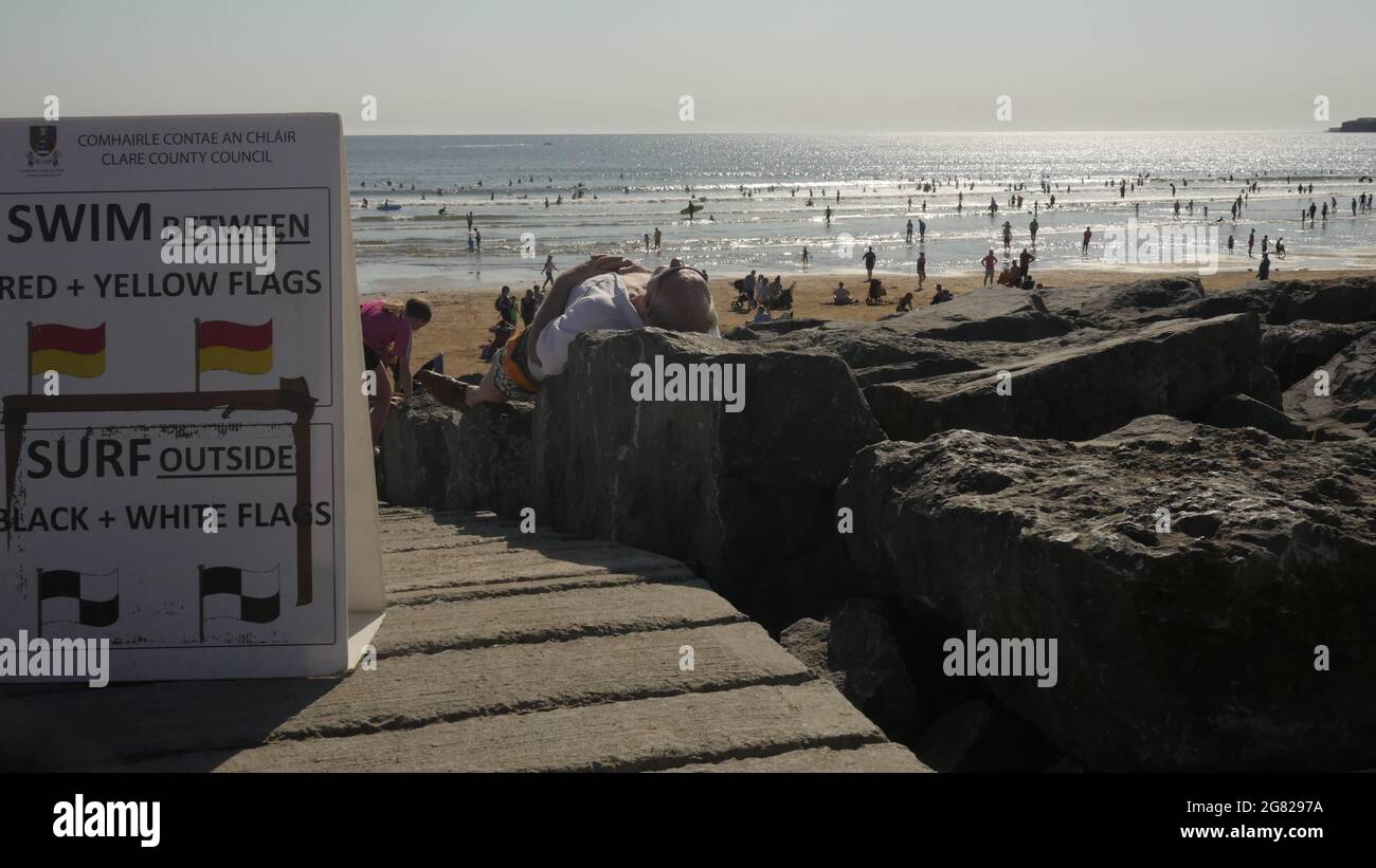 Una spiaggia soleggiata con un sacco di persone in mare e un uomo prendere il sole su alcune rocce. Foto Stock