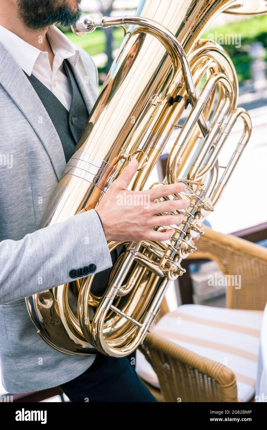 Primo piano delle mani di un musicista jazz che suona una tuba d'oro. Si trova su una terrazza esterna, vestito con una giacca grigia e barba tinta blu scuro. Foto Stock