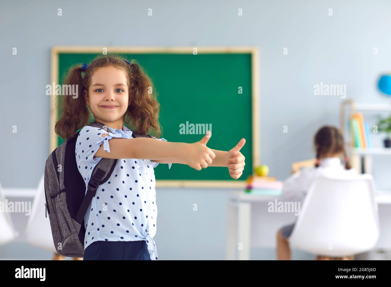 Adorabile bambina con zaino che mostra i pollici in classe. Bambino felice che torna a scuola, puntando al successo Foto Stock