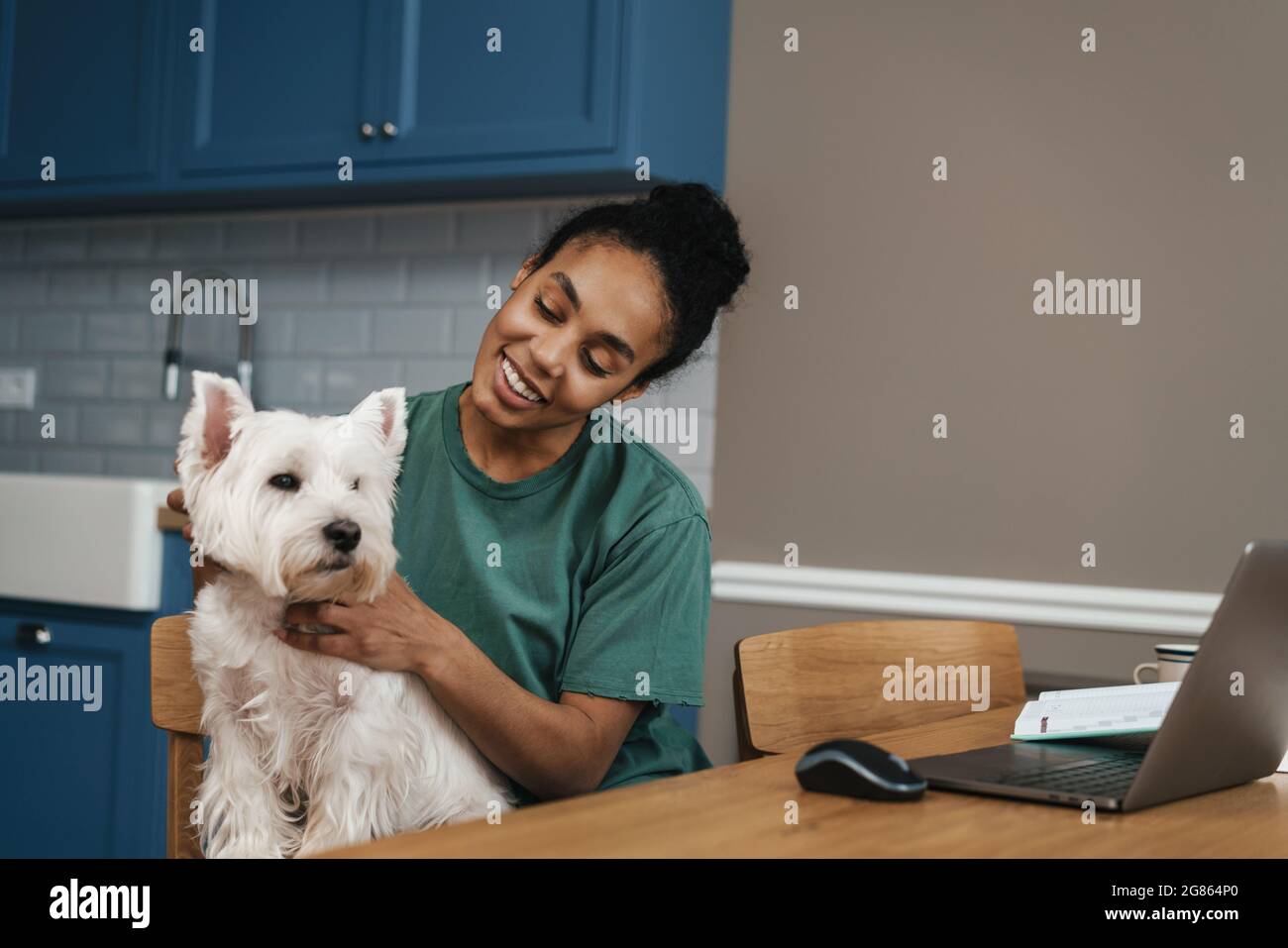 Sorridente donna africana di mezza età seduto al tavolo all'interno con il suo cane in una videochiamata via computer portatile Foto Stock