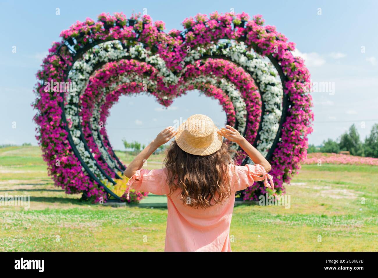 giovane donna in abito rosa e cappello si erge su uno sfondo di archi fioriti. Immagine con una messa a fuoco selettiva sul cappello. Arco nuziale a forma di cuore Foto Stock