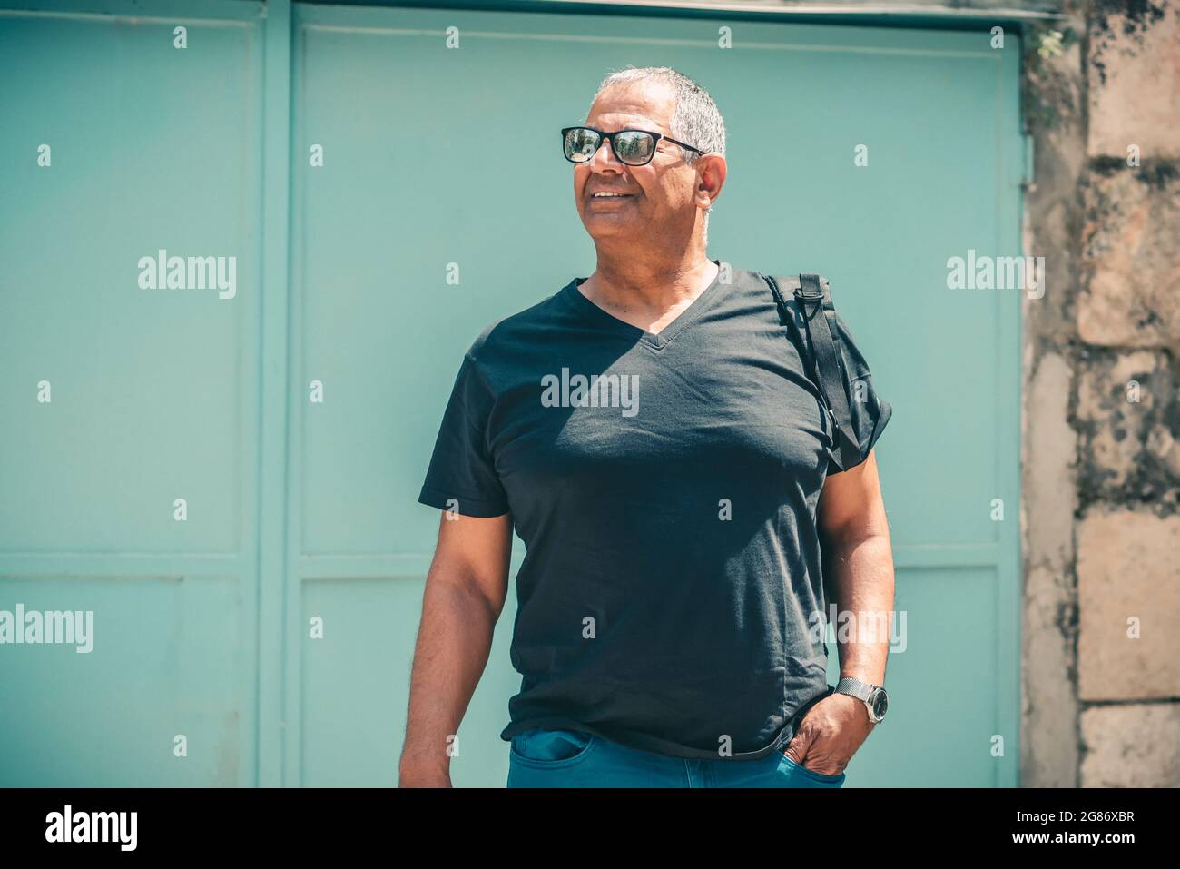 Uomo maturo sorridente che si trova di fronte a un vecchio edificio che tiene il suo zaino nero, guardando da parte. Viaggi di stile senior. Foto Stock