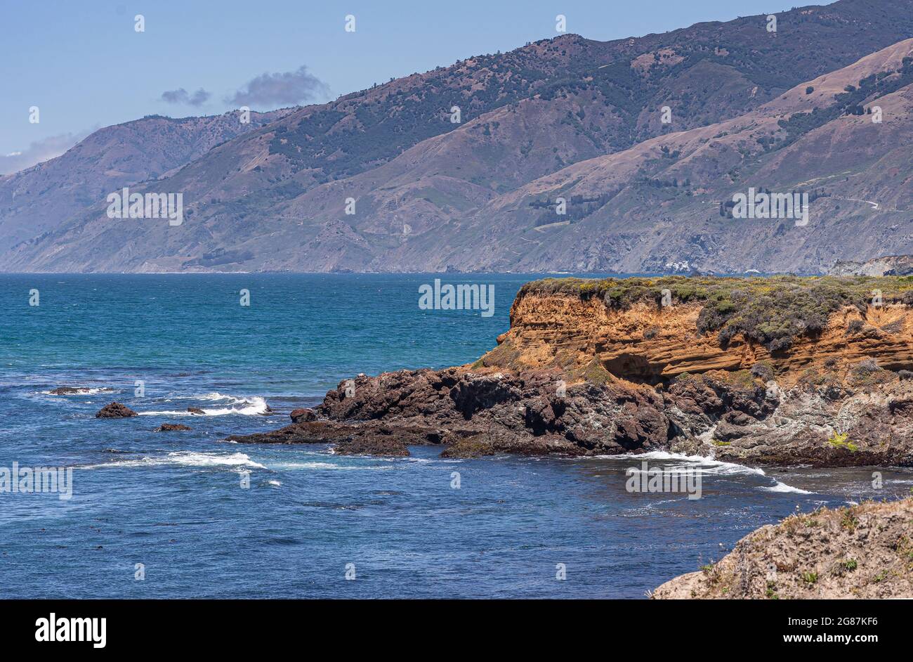 San Simeon, CA, USA - 8 giugno 2021: Costa dell'Oceano Pacifico a nord della città. Scogliere di roccia rossa in acque blu profonde vicino a Ragged Point con Los Padres mounts Foto Stock