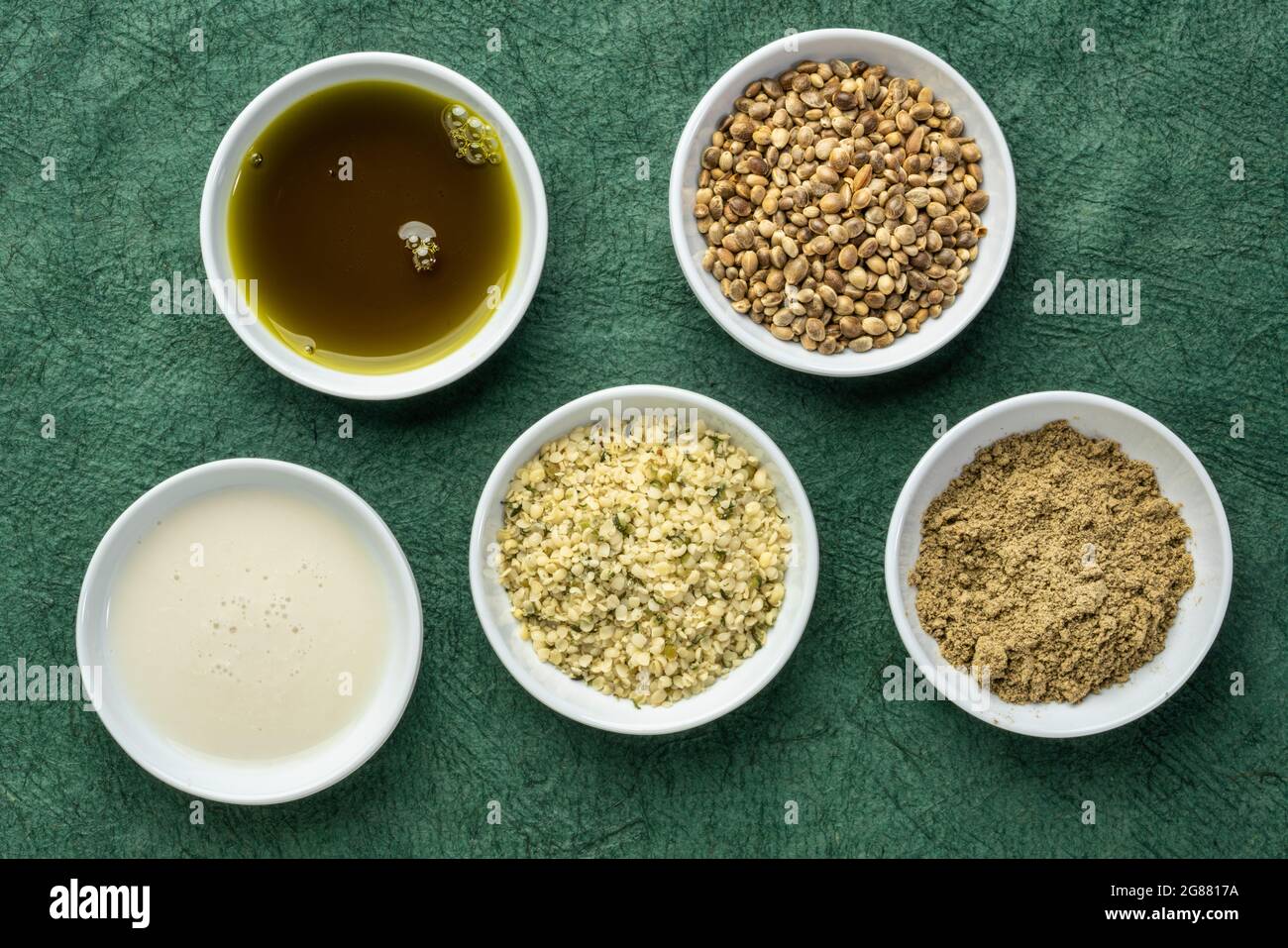 raccolta di prodotti di semi di canapa: cuori, proteine in polvere, latte e olio in piccole ciotole bianche contro carta verde testurizzata, concetto di supercibo Foto Stock