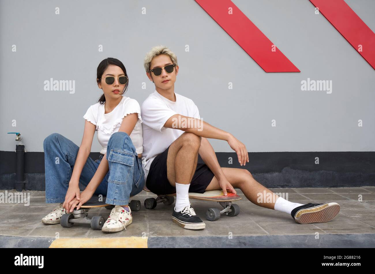 Coppia asiatica che riposa dopo lo skateboard. Giovane asiatico in T-shirt bianca e occhiali da sole neri poggiano il braccio al ginocchio. La sua ragazza seduta accanto Foto Stock