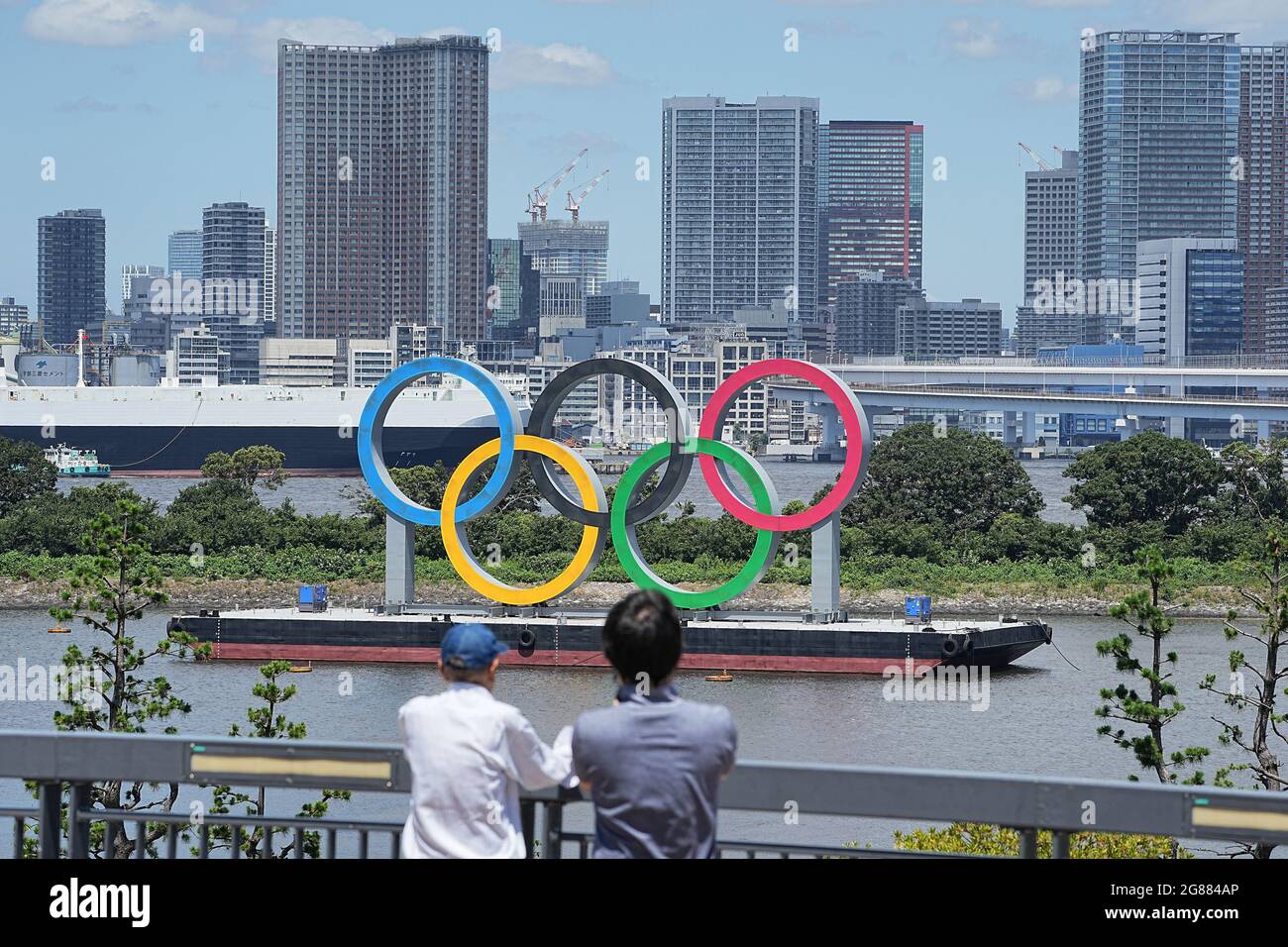 Tokio, Giappone. 18 luglio 2021. Due persone si trovano su una baia di fronte agli Olimpiadi di grandi dimensioni. Gli anelli si ergono su una zattera nella Baia di Tokyo di fronte al quartiere di Odaiba. Le Olimpiadi di Tokyo 2020 si terranno dal 23 luglio 2021 all'8 agosto 2021. Credit: Michael Kappeler/dpa/Alamy Live News Foto Stock