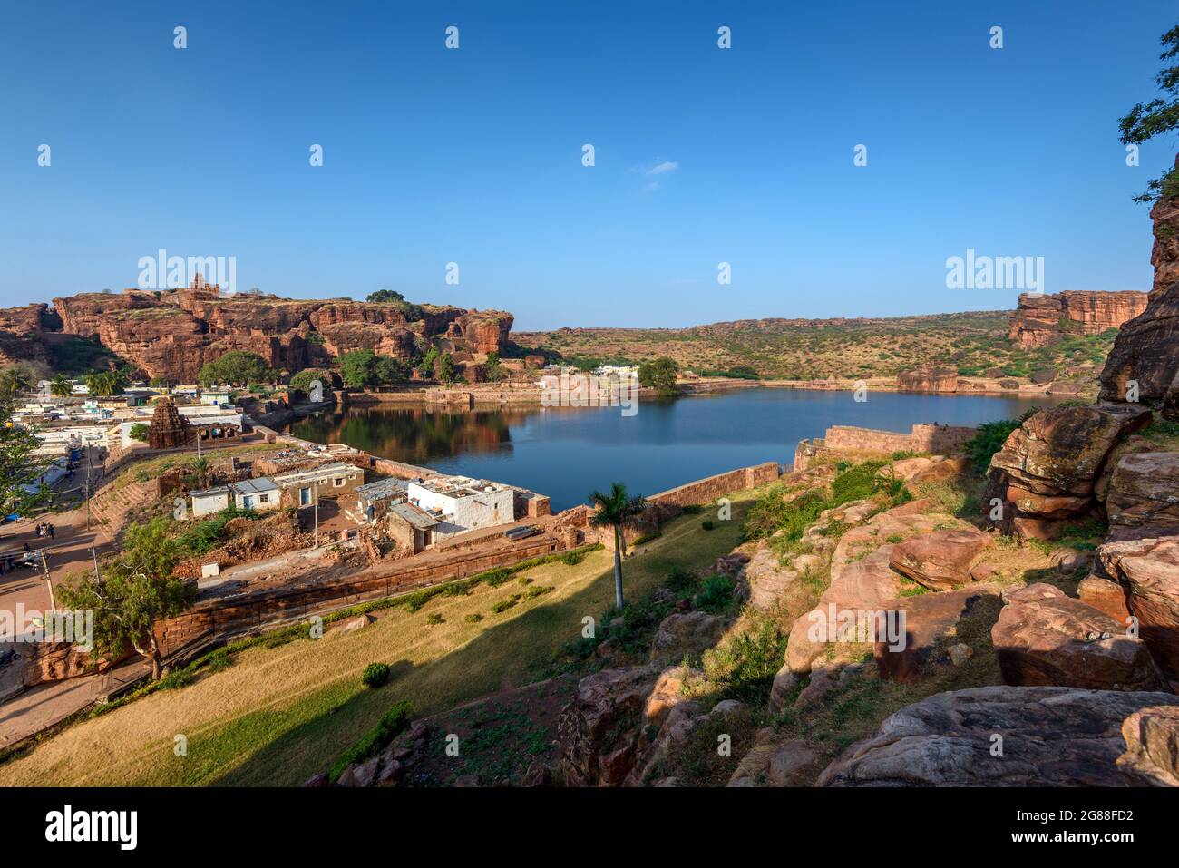 Badami, Karnataka, India - 10 gennaio 2020 : Lago di Agasthya e colline di roccia rossa circostanti. Alcuni edifici in pietra marrone tempio spruzzato intorno, Badami Foto Stock