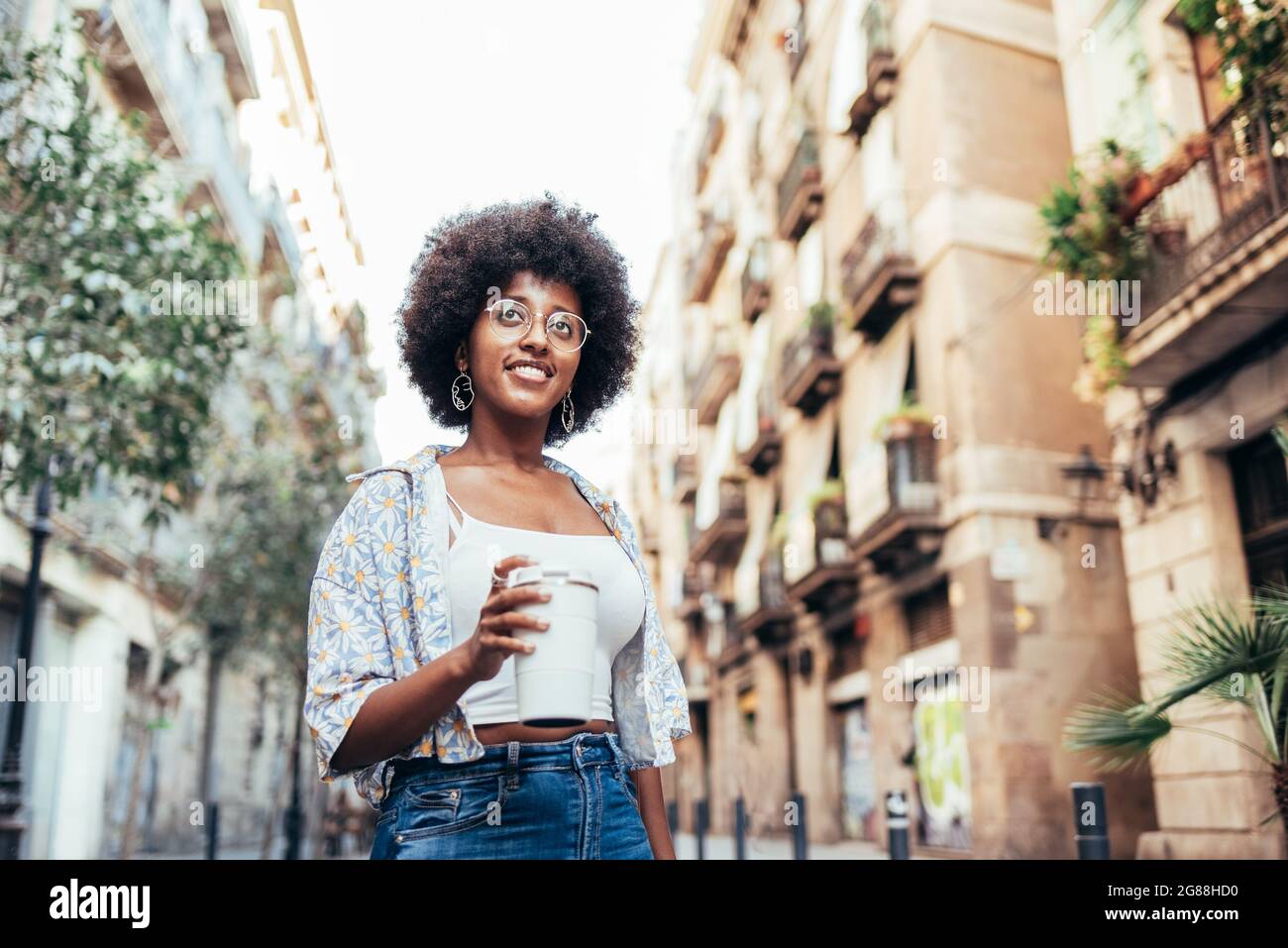 ritratto orizzontale di una giovane donna africana. Sorride e tiene una tazza riutilizzabile di caffè. Concetto di mattina d'estate in città Foto Stock