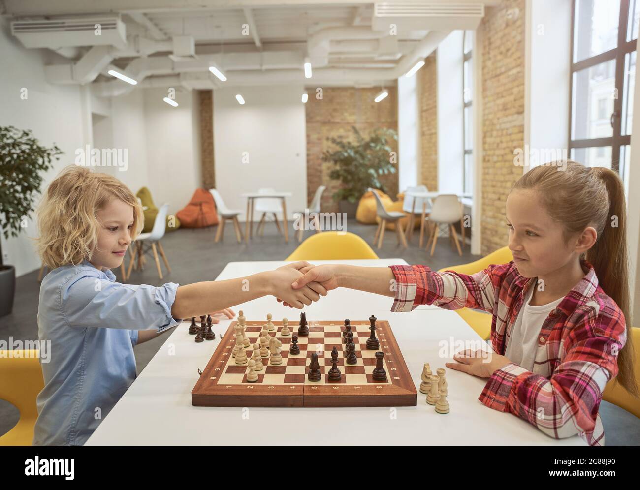 I bambini piccoli intelligenti, il ragazzo e la ragazza che scuotono le mani dopo la partita, giocando a gioco di tavolo, seduti insieme al tavolo a scuola Foto Stock