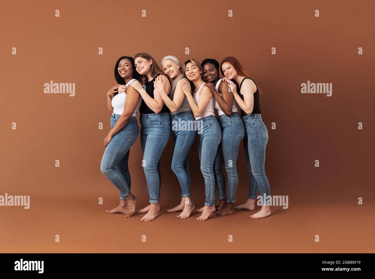 Sei donne sorridenti che si abbracciano in studio. Femmine di diverse età e tipi di corpo che si posano su sfondo marrone. Foto Stock