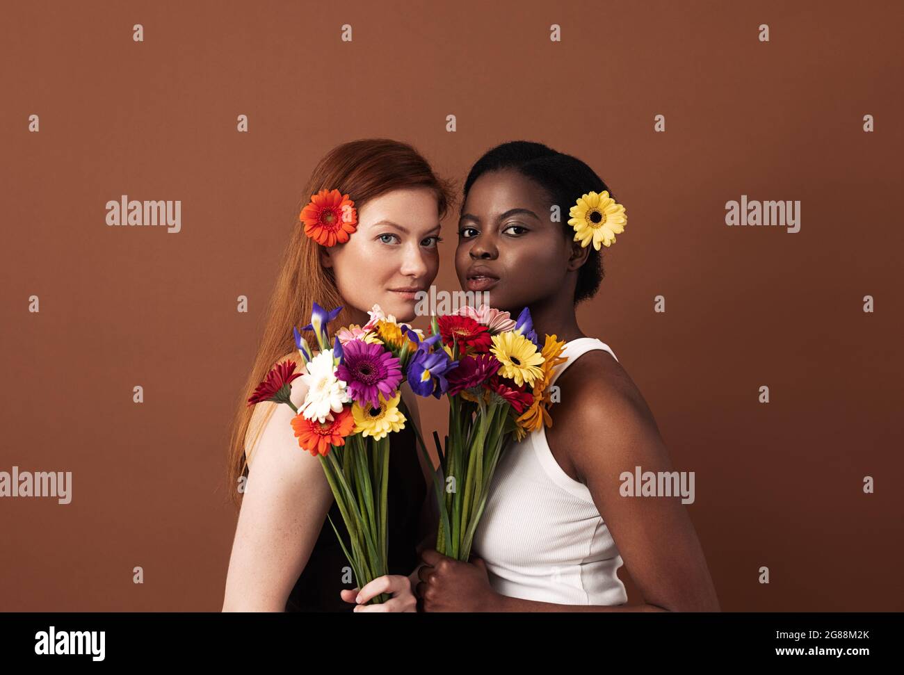 Due donne di razze diverse con fiori nei loro capelli che tengono bouquet. Caucasiche e afroamericane che guardano la macchina fotografica in uno studio. Foto Stock