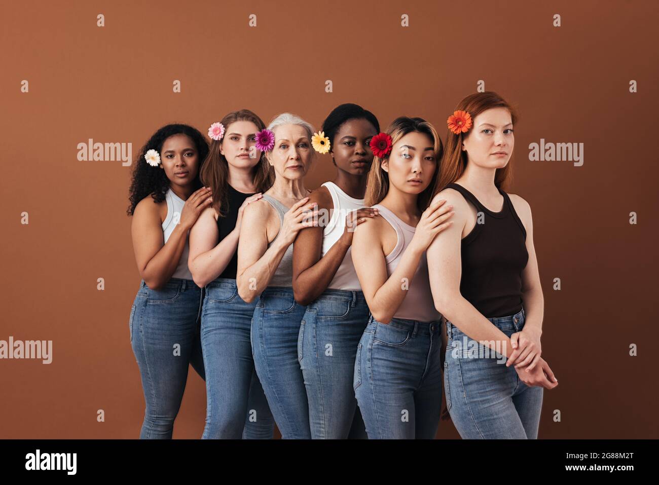 Sei donne diverse che si levano insieme su sfondo marrone. Donne di età diversa con fiori nei capelli. Foto Stock