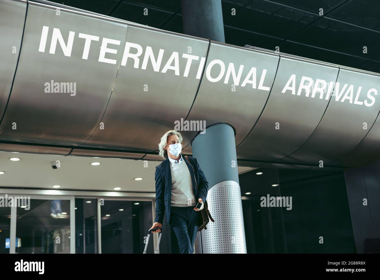 Uomo d'affari che esce dal cancello degli arrivi internazionali all'aeroporto. Uomo che indossa la maschera al terminal dell'aeroporto dopo la pandemia. Foto Stock