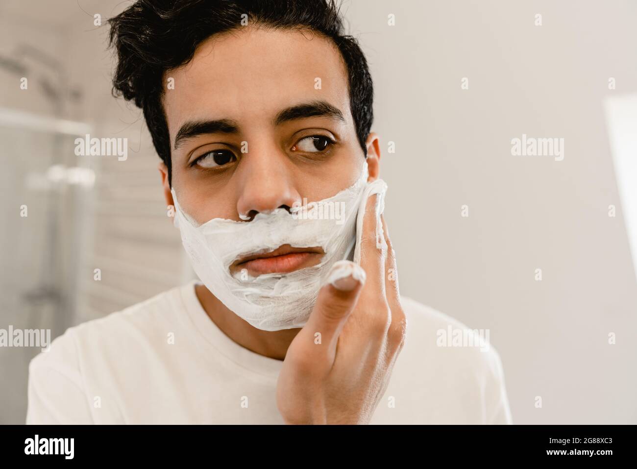 Giovane uomo ispanico che si rade il viso in piedi allo specchio all'interno, primo piano Foto Stock