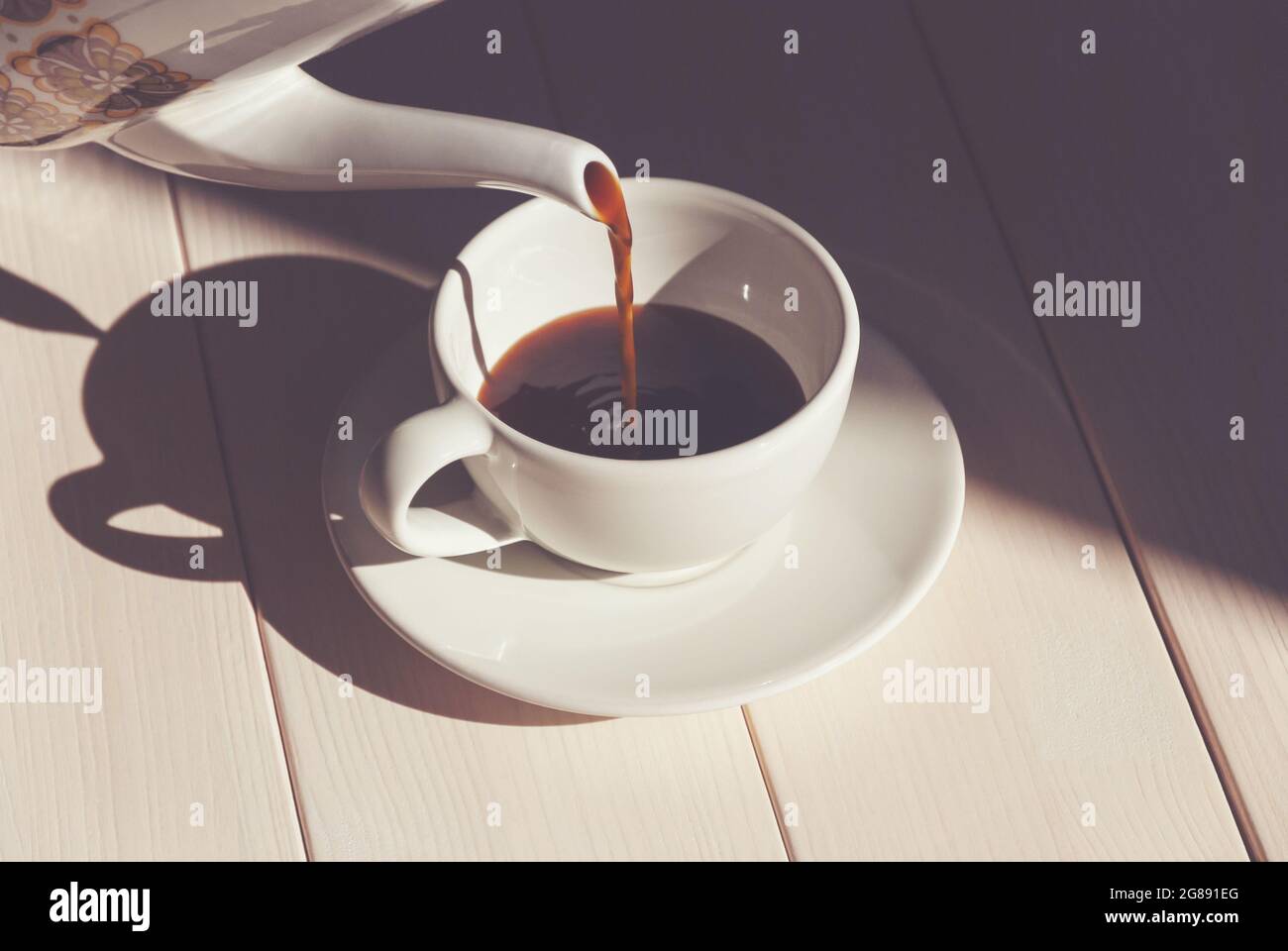 Versare il caffè nella tazza - mattina estiva con luce solare e ombre luminose, caffettiera, tazza bianca e piattino, serve caffè Foto Stock