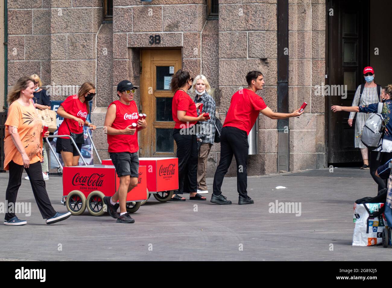 Persone in camicie rosse Coca-Cola che consegnano lattine di cola ai passanti in una giornata calda alla stazione ferroviaria centrale di Helsinki, Finlandia Foto Stock