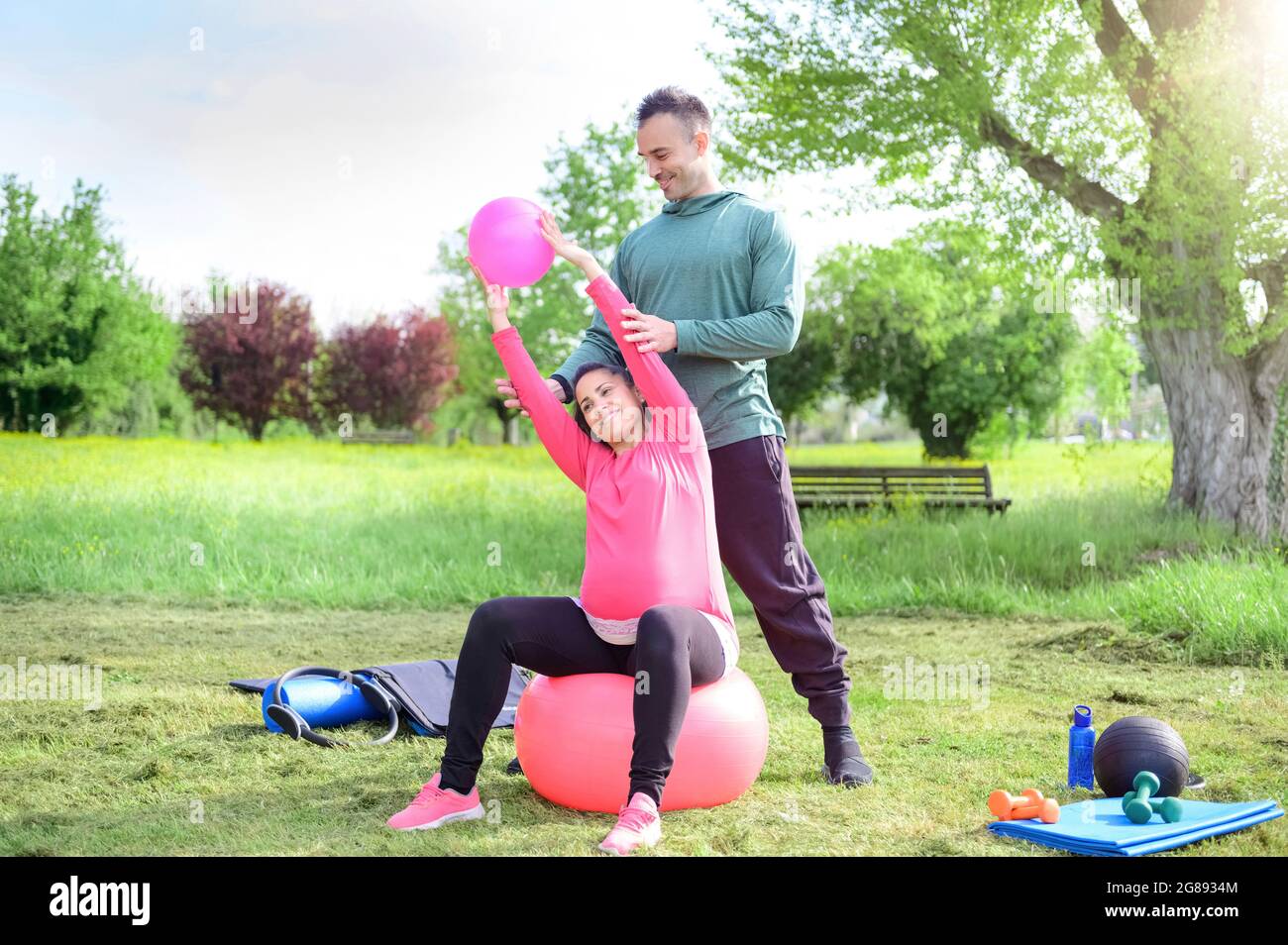 Sorridente personal trainer e una donna in gravidanza felice facendo yoga esercizio all'aperto in un parco - la ragazza fa muscolo si allunga seduta sulla palla palestra - concetto A. Foto Stock