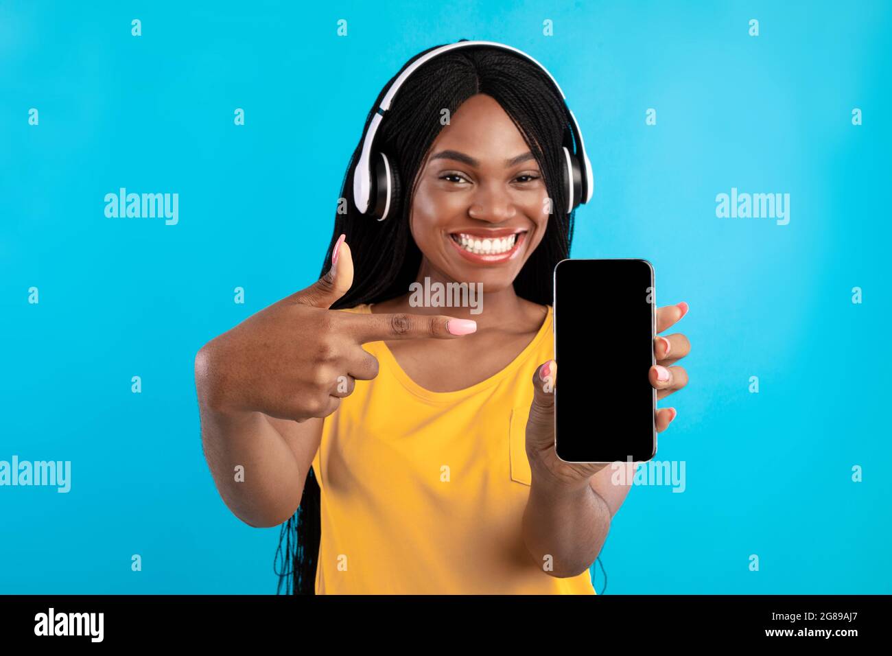 Donna millenaria nera che mostra schermo del telefono indossando cuffie, sfondo blu Foto Stock