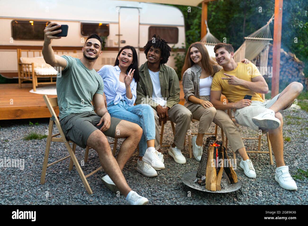 Diversi giovani amici che prendono selfie vicino al fuoco di sera, utilizzando smartphone, seduti accanto al camper durante il viaggio in campeggio Foto Stock