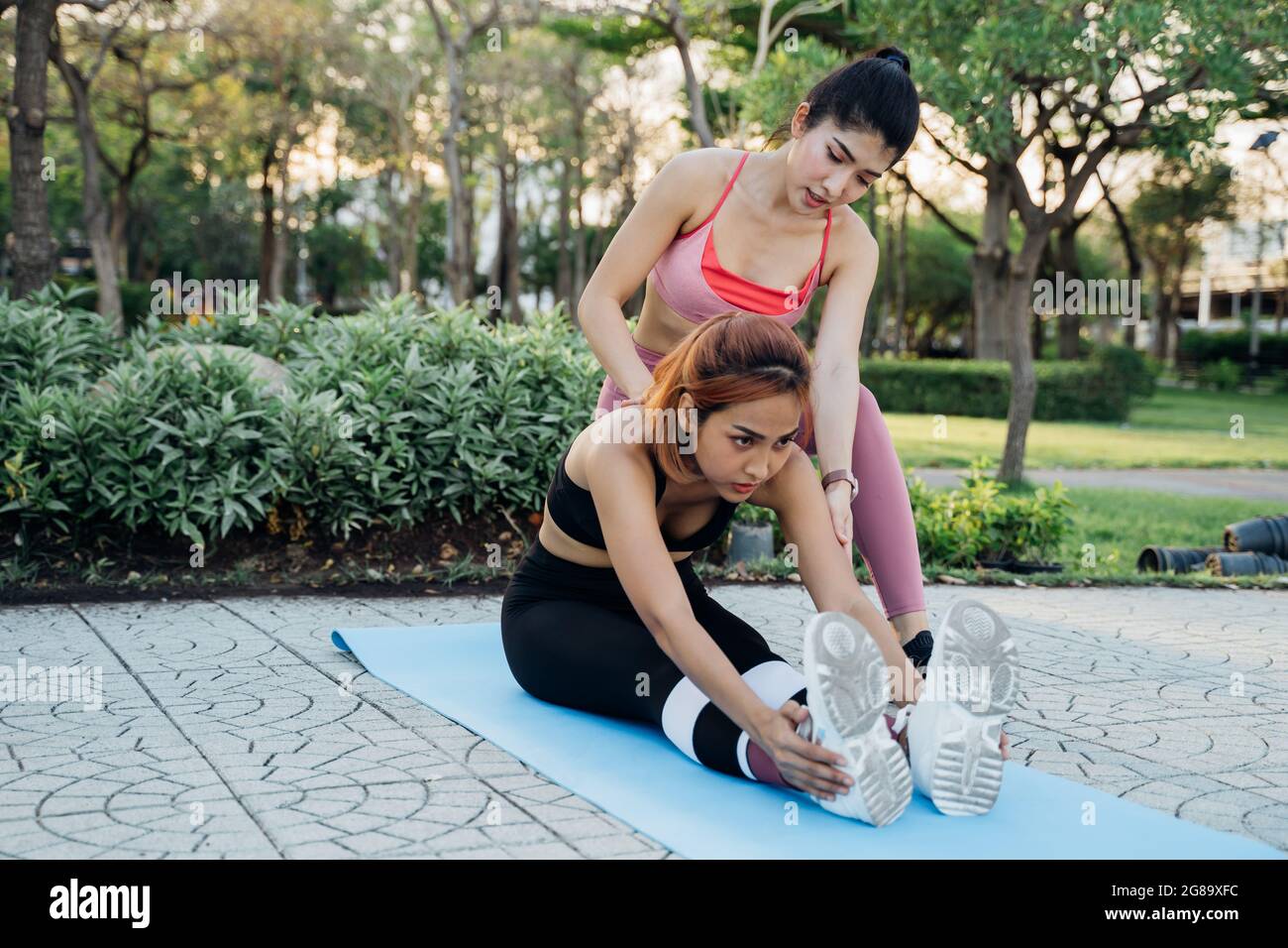 Giovane donna asiatica che tiene le gambe della sua amica femminile seduta sul tappeto di yoga che pratica l'esercizio di stretching nel mornging nel parco Foto Stock