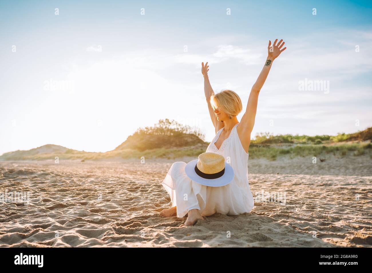 Donna bionda che indossa lunghi abiti bianchi siediti sulla spiaggia vuota  e guarda il mare con le mani in su. Libertà, felicità Foto stock - Alamy