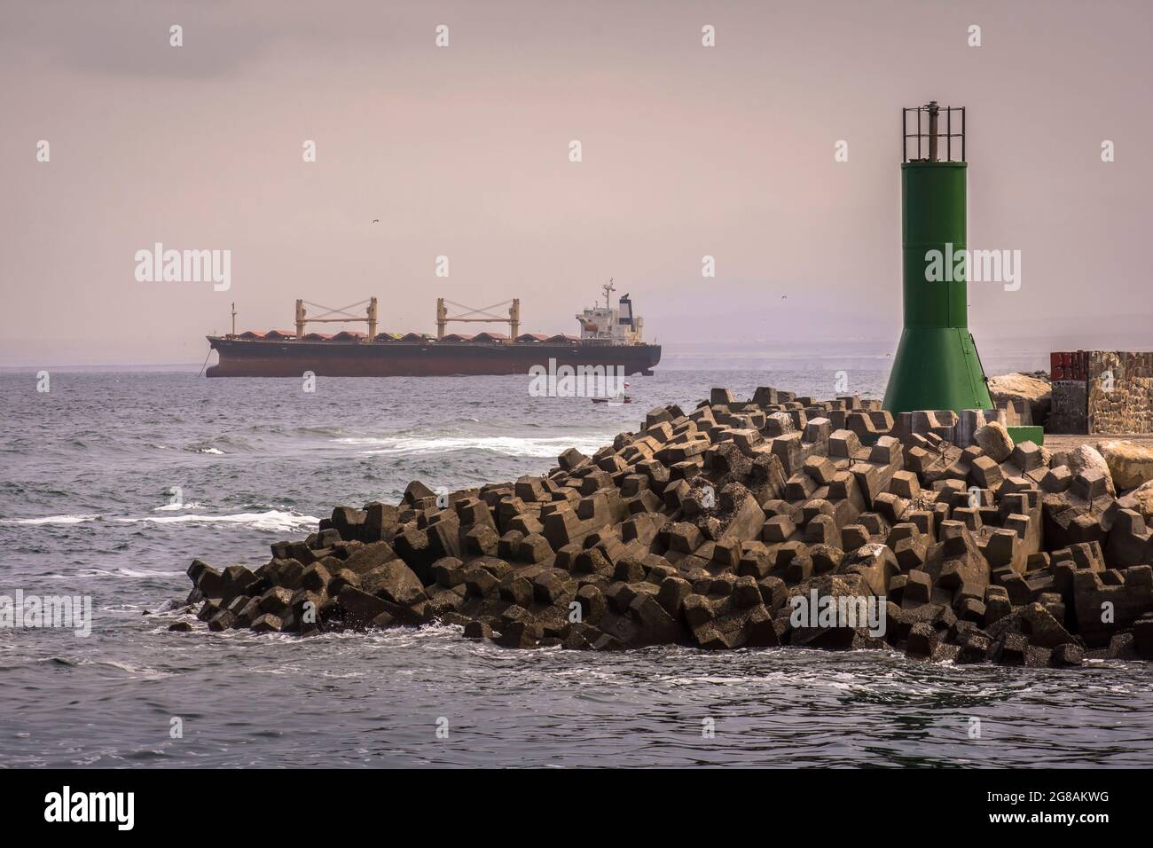 Frangiflutti fatti con cubetti di cemento e una torre verde con una piccola barca e una grande nave in mare. Antofagasta, Cile Foto Stock