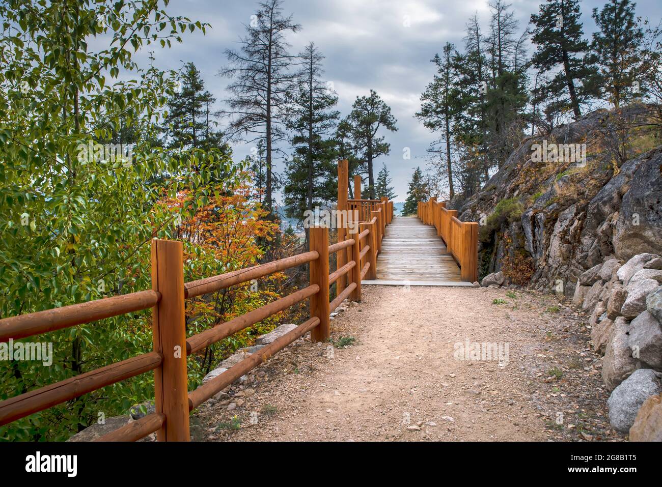 strada sterrata per un ponte di legno con un recinto di tronchi vicino a una scogliera di pietra tra gli alberi. Cielo blu con nuvole sullo sfondo. Foto Stock