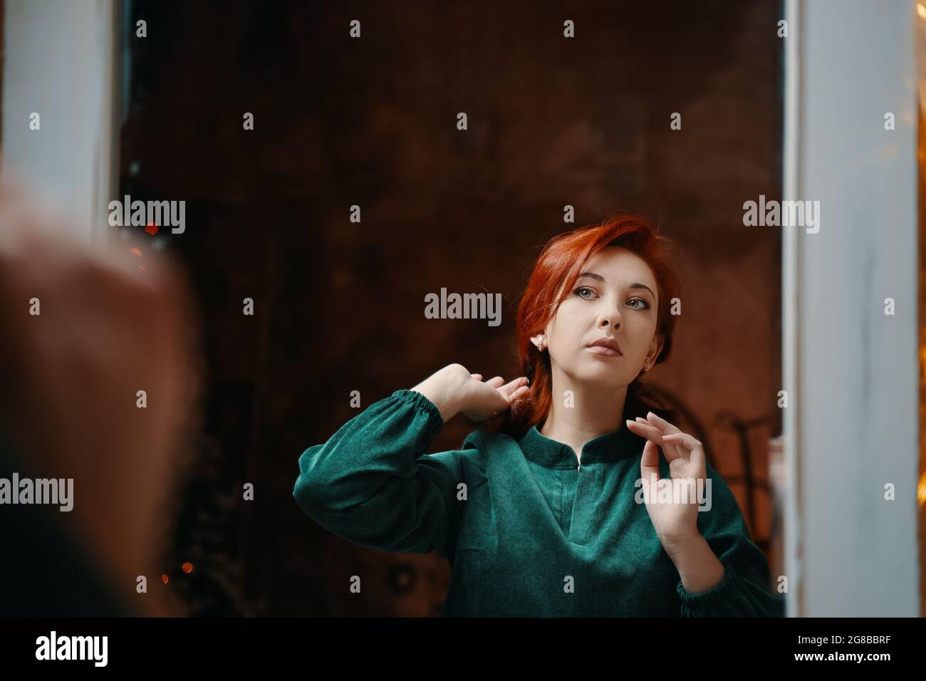 Nobile ragazza dai capelli rossi si ammira nello specchio. Favolosa ragazza retrò in velluto vestito verde si siede e regola il suo coiffure. Parete marrone vintage su sfondo. Foto Stock