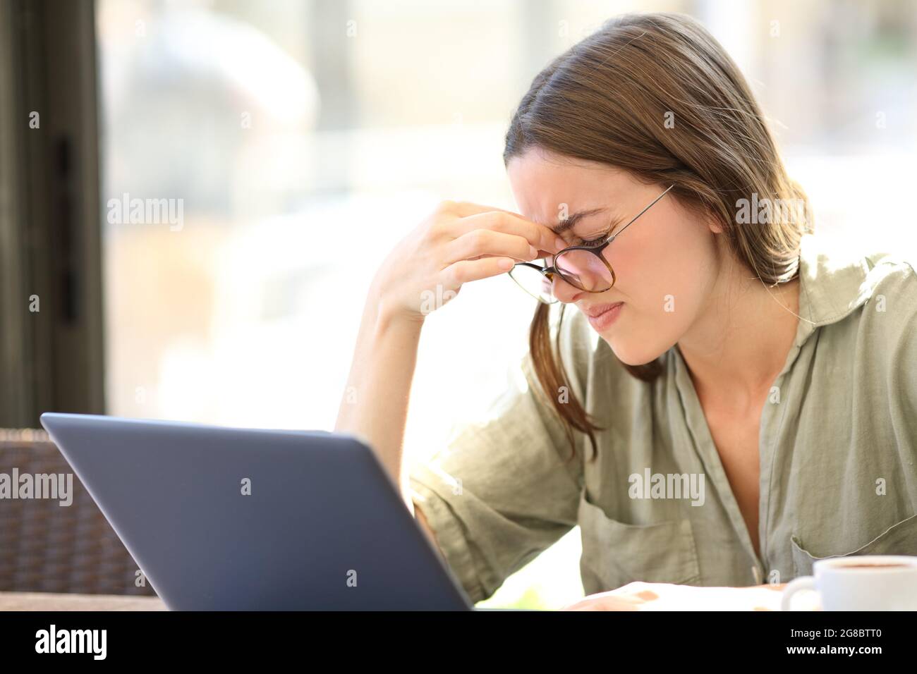 Donna stanca che indossa occhiali che soffrono di pioggia battente usando un computer portatile seduto in un bar Foto Stock