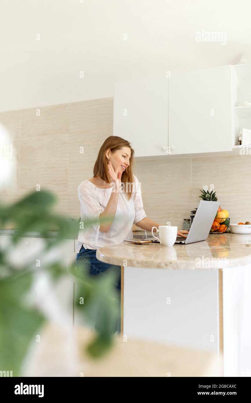 Ritratto di una ragazza bellissima e sorridente che usa un computer portatile sul posto di lavoro. Scuotendo la mano per dire Ciao qualcuno dal suo laptop. Incontri online o soggiorno Foto Stock