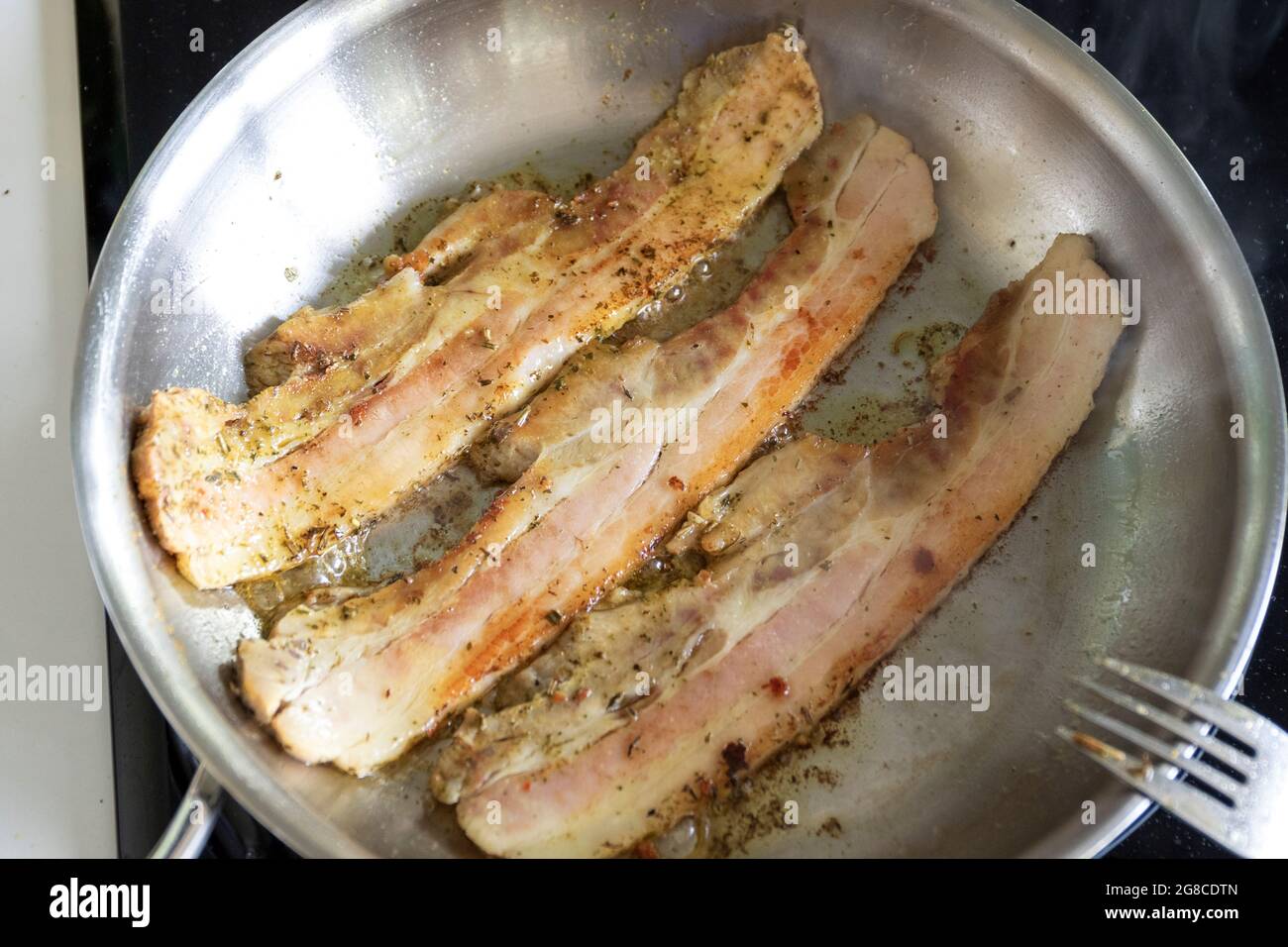 Un ritratto dall'alto di alcuni pezzi di pancetta di maiale adagiati in una  padella in acciaio inossidabile con una forchetta laterale. La carne è  sfrigolante nella padella e così Foto stock -