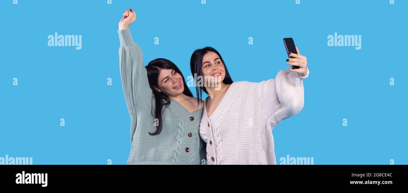 Ragazze gemelle allegre che fanno selfie isolato su sfondo blu una di loro ha alzato la mano destra in su e sorridente. Foto Stock