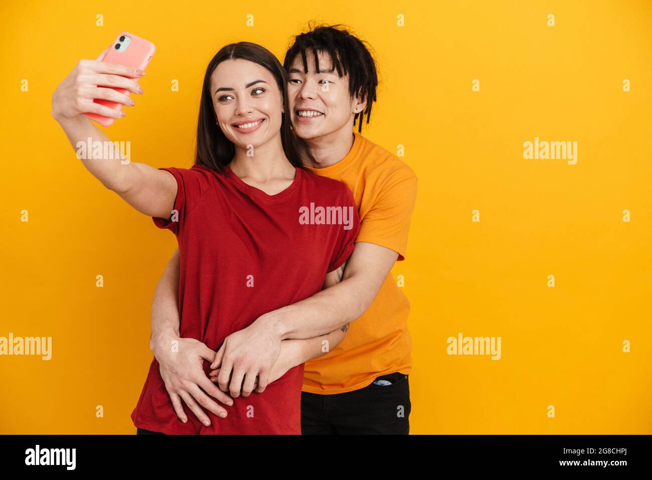 Felice giovane coppia multietnica su sfondo giallo, prendendo un selfie, abbracciando Foto Stock