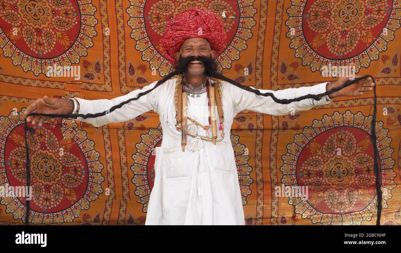 Vecchio maschio indiano che indossa abiti bianchi e un turbante rosso con un volto felice che tiene i suoi lunghi baffi Foto Stock