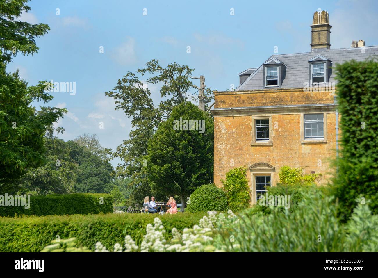 La gente glamour siede sul patio davanti ad una bella villa georgiana chiamata Hadspen House nel Somerset. Fa parte del nuovo Newt Hotel vicino a Bruton. Un lussureggiante giardino si trova in primo piano. L'immagine riflette un classico, bella casa di campagna inglese con la gente che la gode sotto il sole. Foto Stock