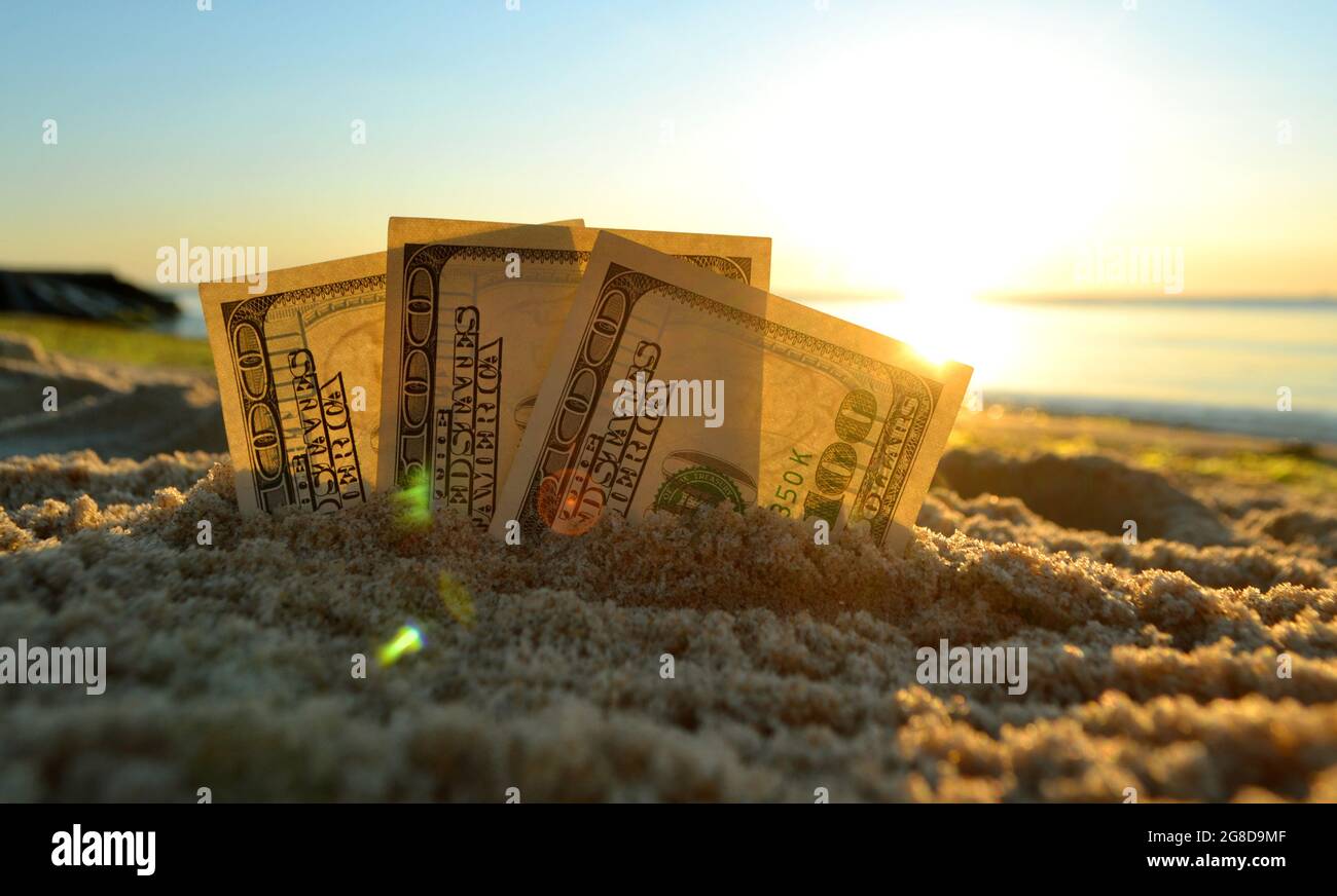 Tre dollari di bollette sono sepolti in sabbia sulla spiaggia di sabbia vicino al mare al tramonto Foto Stock