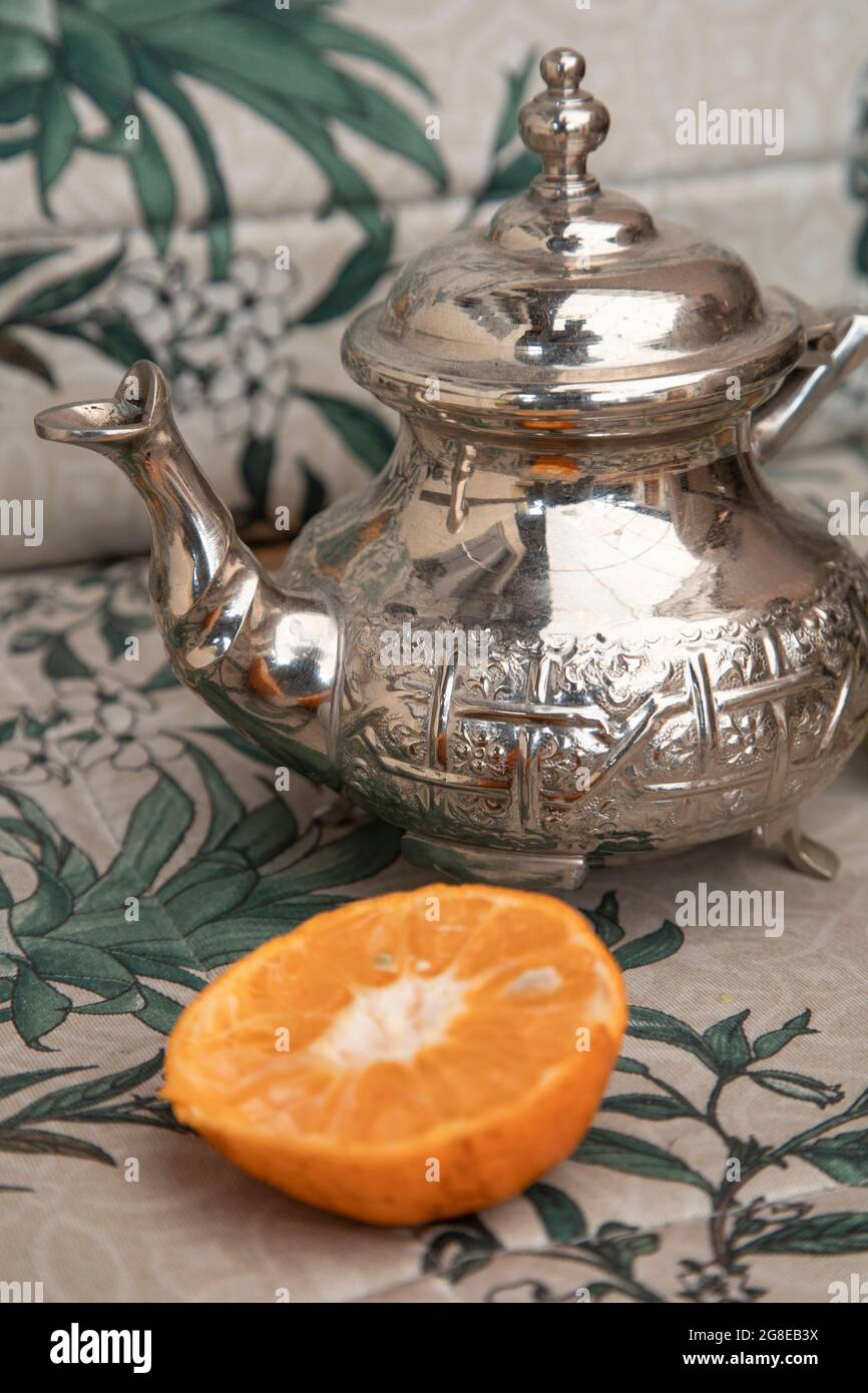 teiera tradizionale in metallo accanto ad una fetta arancione, sullo sfondo una tovaglia con un motivo di foglie, studio con oggetti da cucina Foto Stock