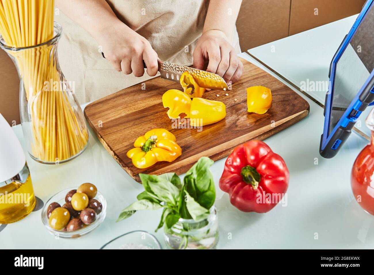 Teenager prepara un seminario virtuale online, taglia il pepe giallo, guarda la ricetta digitale sul tablet touchscreen mentre prepara un pasto sano nel Foto Stock