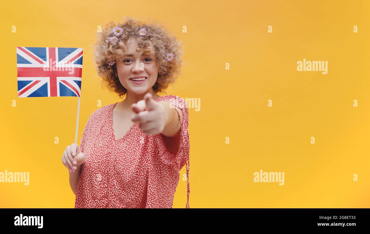 Ragazza che tiene in mano la bandiera britannica. Puntare con il dito verso la fotocamera. Ragazza isolato su uno sfondo giallo con la Gran Bretagna Bandiera. Imparare l'inglese concetti. Foto Stock