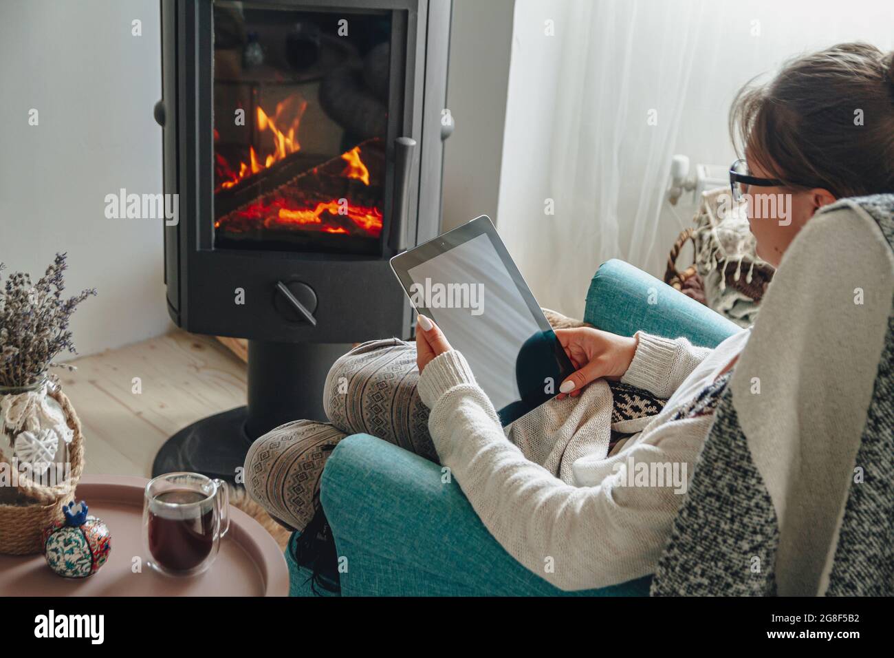 donna accanto al camino, seduta in una comoda poltrona, con un caldo Foto Stock