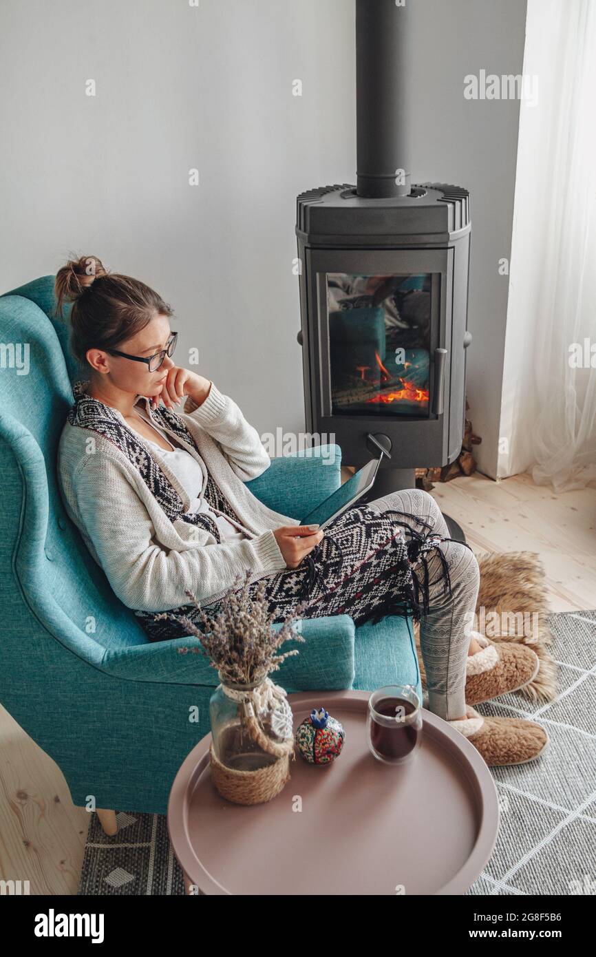 donna accanto al camino, seduta in una comoda poltrona, con un caldo Foto Stock