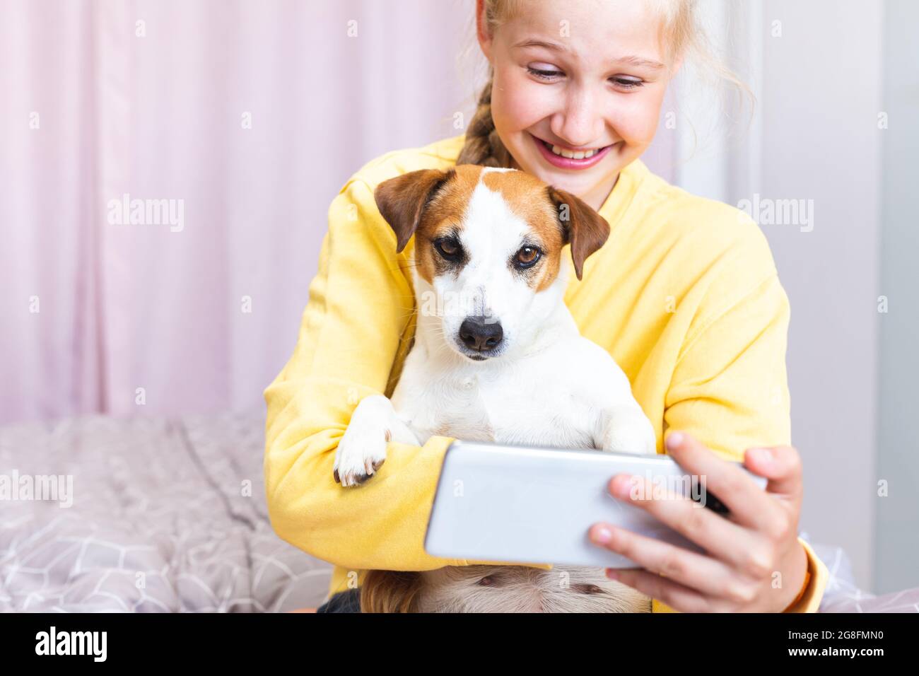 Una ragazza ridente con un telefono cellulare in mano, insieme al suo cane Jack Russell Terrier, comunica sui social media o via Zoom, fa un selfie Foto Stock